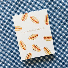Load image into Gallery viewer, A Weenie Bit Older Card, hand-painted watercolor hot dog pattern with mustard and gold foil accents on textured white card, on blue and white patterned fabric surface. 