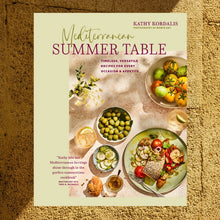 Load image into Gallery viewer, Mediterranean Summer Table Cookbook, cream-colored cookbook cover featuring the title "Mediterranean Summer Table" by Kathy Kordalis above a styled Mediterranean meal scene with bowls of green olives, sliced cucumbers, and vine tomatoes, alongside lemon drinks, crusty bread, and a plated fish entrée on a soft pink stone tabletop, against a textured tan  background. 