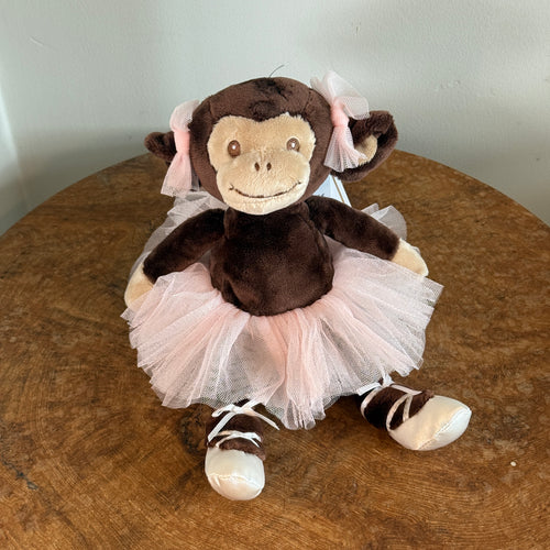 Plush monkey with dark brown fur, light beige face and hands, stitched brown smile and eyes, wearing a pink tulle tutu with matching pink bows on its ears and white satin ballet-style shoes with ribbon laces. Monkey is sitting upright on a wooden surface and against a white background.  