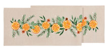 Load image into Gallery viewer, Cedar and Citrus Table Runner, natural beige fabric table runner folded to show its horizontal garland of orange slices, green leaves, evergreen sprigs, brown star anise, and small white dots, against a white background. 