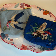 Load image into Gallery viewer, Light blue velvet mini pouch embroidered with a blue and white swallow detailed with gold sequins and beads, carrying pink flowers, next to a royal blue velvet mini pouch embroidered with a brown, gold, and white owl detailed with gold sequins. The owl is perched on a wood stump among red and white mushrooms and green ferns. Both pouches are lying side by side on a white scalloped dish, with a red, orange, green, and brown botanical design. 