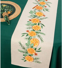 Load image into Gallery viewer, Cedar and Citrus Table Runner, natural beige fabric runner with a central garland design of orange slices, green leaves, evergreen sprigs, tan star anise,  and small white dots, displayed on an emerald green tablecloth beside a green napkin in a woven napkin ring and a round woven placemat with a dark green center. 