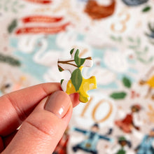 Load image into Gallery viewer, Close-up of a hand holding a single illustrated puzzle piece above the 12 Days of Christmas jigsaw puzzle in a soft focus.