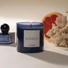 Load image into Gallery viewer, Poured NY Bicoastal candle in blue glass vessel, displayed lit on a white surface with coordinating blue Bicoastal parfum  bottle, citrus slice, and coral decor in the background.
