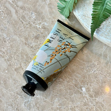 Load image into Gallery viewer, Fresh Sea Salt Demi Hand Cream, pale blue tube with orange, yellow, and white botanical accents, a black cap, and black crimped end displayed beside green fern fronds on a beige marble surface. 
