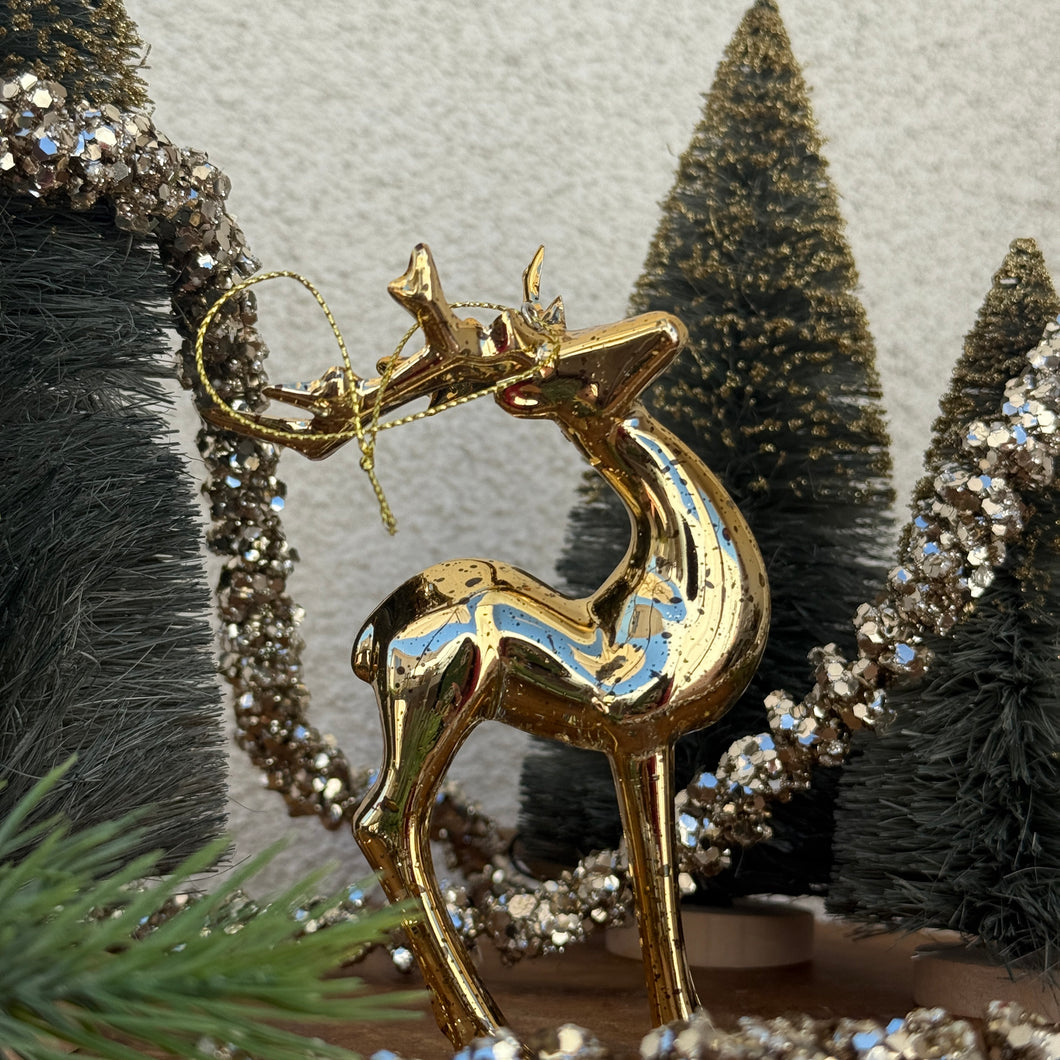 A gold metallic deer ornament with a gold hanging string looped around the antlers. Three green decorative Christmas trees, a green fir tree cutting, and a strand of silver glitter garland behind the deer. On top of a wooden surface. Against a white textured background. 