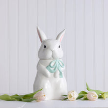 Load image into Gallery viewer, White ceramic rabbit cookie jar with removable head lid and soft green bow, styled on white surface with pale pink tulip flowers.