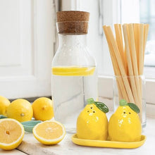 Load image into Gallery viewer, Lemon Salt &amp; Pepper Shakers, glossy yellow ceramic lemon-shaped salt and pepper shakers with light green stems and dark green leaves, displayed on a matching yellow ceramic oblong tray beside a glass carafe with a brown cork stopper, water, and a lemon slice, a clear glass of wooden utensils, and fresh lemons on a light stone surface. 