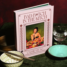Load image into Gallery viewer, Bad B*tch In The Kitchen by Cassie Yeung, pale pink hardcover cookbook with portrait cover illustration, red geometric border details, and floral corner motifs displayed beside patterned bowl with chopsticks, turquoise dish, and cut-glass bowl on dark green fabric surface against a deep red background.