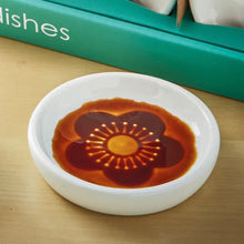 Load image into Gallery viewer, Secret Sauce Dish round white ceramic sauce dish with single embossed flower design revealed by sauce, shown on a surface with packaging in background.  
