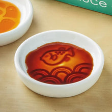 Load image into Gallery viewer, Secret Sauce Dish round white ceramic sauce dish with single embossed fish and wave design revealed by sauce, shown on light surface with packaging in the background. 