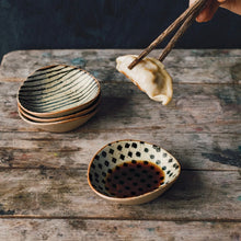 Load image into Gallery viewer, Unite Element Dipping Dishes, set of four, stoneware dishes with reactive glaze and black patterns (stripe and dot) on cream speckled interiors, beige textured exteriors, and tan rims, shown with sauce and dumpling on a rustic wood surface.  