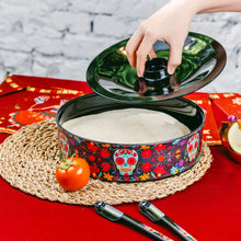 Load image into Gallery viewer, Day of the Dead Tortilla Holder, round black tortilla warmer with aqua sugar skull and colorful floral design, hand lifting lid to reveal tortillas inside, and matching black tongs displayed on red surface.  
