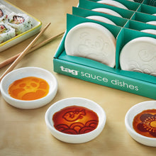 Load image into Gallery viewer, Secret Sauce Dish round white ceramic sauce dishes with hidden embossed panda, fish, and flower designs revealed by sauces, shown with packaging on a light surface with chopsticks and sushi rolls in background. 
