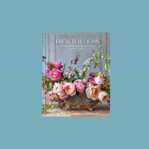 Book cover of French Blooms by Sandra Sigman featuring a floral arrangement of pink and peach colored roses with greenery displayed in a decorative vessel against a soft gray background with white lettering. Book against a light blue background. 