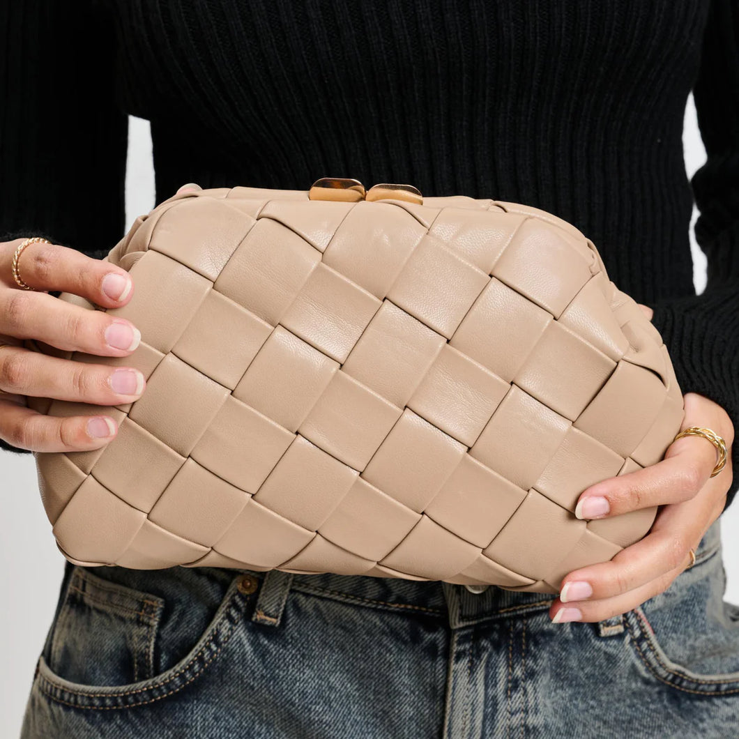 Model holding a woven beige vegan-leather crossbody bag with a structured half-moon shape and gold clasp detail, styled with denim jeans and black ribbed sweater.
