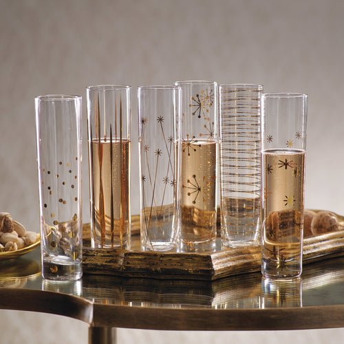 Assorted clear glass champagne flutes with gold decals on a gold tray with a glass top, with champagne poured into three of six flutes.