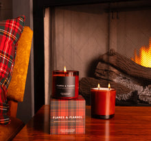 Load image into Gallery viewer, Two lit &quot;Flames &amp; Flannel&quot; candles in red glass jars on a wooden table, one featuring a black label and resting on a red plaid gift box, the other without a label placed beside a glowing fireplace. A red and green plaid pillow and a golden chair appear on the left side. 