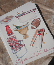 Load image into Gallery viewer, White illustrated print featuring hand-drawn watercolor art of tailgate essentials, including hot dogs, bottles in tubs, red cups, a folding lawn chair, a football, and a cornhole board, with "Tailgates and Touchdowns" in red cursive text at the bottom, shown resting on a brown surface. 