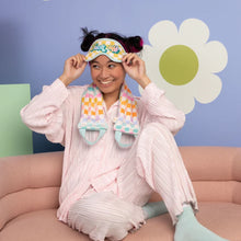 Load image into Gallery viewer, Person in light pink loungewear and blue socks sitting on a pink sofa wearing a rainbow-checkered weighted neck wrap in blue, lavender, orange, pink, and white squares, and holding a matching eye mask on their head against a light purple background featuring a white retro daisy with a lime green center.   