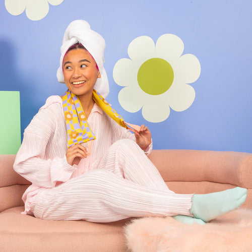 Smiling person in light pink loungewear and blue socks sitting on a pink sofa with a white towel wrapped around their head, wearing a yellow and lavender checkered weighted neck wrap with orange flower accents, set against a light purple background featuring retro white daisies with lime green centers. 
