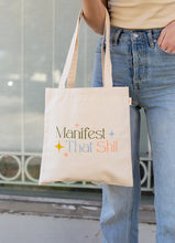 Load image into Gallery viewer, Person wearing a beige top and blue jeans holding a white canvas tote bag with the phrase &quot;Manifest That Shit&quot; printed in olive, blue, and pink colors with blue, pink, and yellow stars, standing outdoors against a light gray iron fence. 