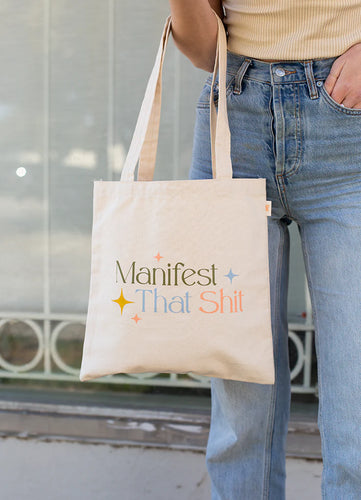 Person wearing a beige top and blue jeans holding a white canvas tote bag with the phrase 