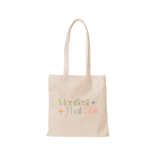 Load image into Gallery viewer, White canvas tote bag with the phrase &quot;Manifest That Shit&quot; printed in olive, blue, and pink colors with pink, blue, and yellow stars, displayed against a black and gray checkered background. 