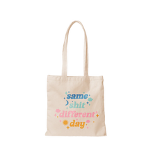 Load image into Gallery viewer, White canvas tote bag with long handles featuring the phrase &quot;same shit different day&quot; in blue, light blue, pink, and orange lettering accented with tiny clouds, sparkles, a sun, and a crescent moon. Displayed against a black and gray checkered background. 