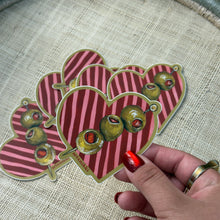 Load image into Gallery viewer, Hand holding heart-shaped stickers with pink-and-red stripes and green olive illustrations, displayed on a tan fabric surface. 