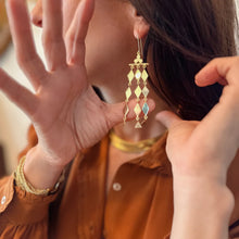 Load image into Gallery viewer, Model wearing the gold-plated brass Kiva statement earrings, showing their long geometric diamond-shaped drops that shimmer and reflect light. 