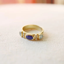 Load image into Gallery viewer, Double-gold plated brass ring featuring a central blue lapis stone flanked by two pale chalcedony stones and accented with small CZ stones, displayed on a textured cream surface. 
