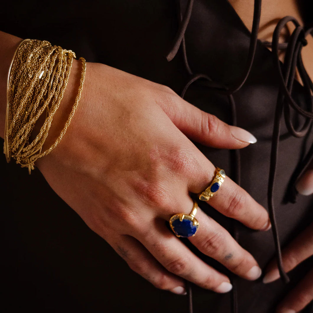 Model's hand adorned with a gold plated multi-strand bracelet and two gold-plated rings with blue lapis stones, resting against a dark brown top featuring thin tie details.  