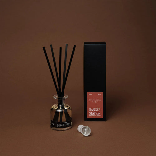 Ranger Station Cowboy Christmas reed diffuser in a reflective glass bottle with black reeds, displayed besides its black box with a red label reading 