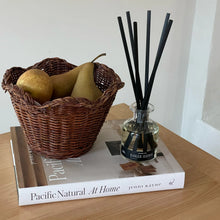 Load image into Gallery viewer, Ranger Station Cowboy Christmas reed diffuser in a reflective glass bottle with black reeds displayed beside a woven basket filled with pears on top of stacked coffee table book titled Pacific Natural: At Home by Jenni Kayne, set on a wooden surface against a white wall. 