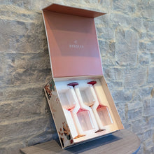 Load image into Gallery viewer, Open Byrdeen gift box with a mauve-colored lid displaying four hand-blown crystal wine glasses with pink gradient stems and bases, set against  a textured gray stone wall on a wooden table  