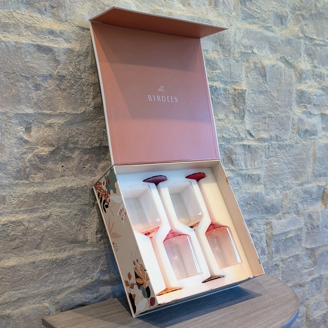 Open Byrdeen gift box with a mauve-colored lid displaying four hand-blown crystal wine glasses with pink gradient stems and bases, set against  a textured gray stone wall on a wooden table  