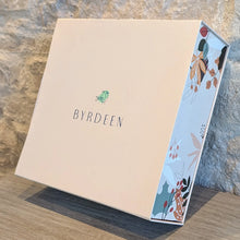 Load image into Gallery viewer, Closed Byrdeen gift box with a cream-colored lid and pastel abstract-patterned sides displayed on a wooden surface against a textured gray stone wall. 