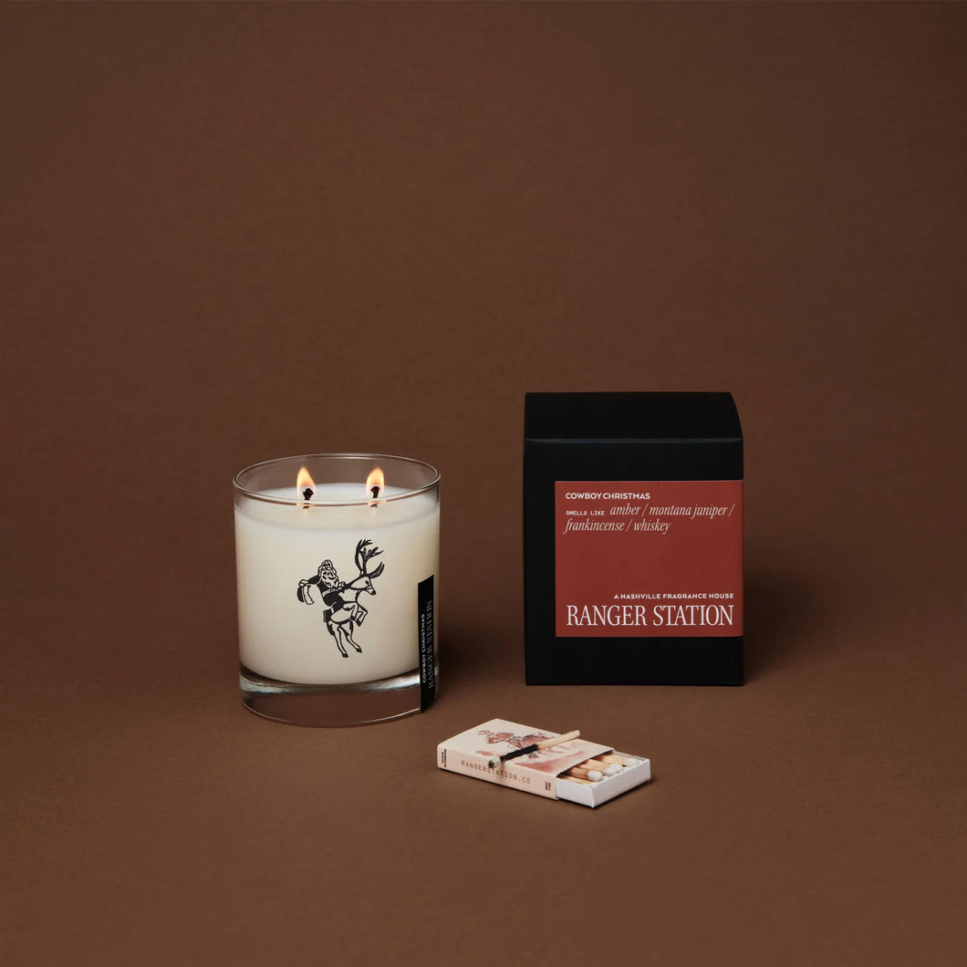 Ranger Station Cowboy Christmas candle with a lit double wick in a clear glass jar featuring a black cowboy illustration, displayed beside its black box with a red label reading 