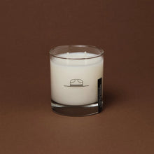 Load image into Gallery viewer, Ranger Station Cowboy Christmas candle with double wicks in a clear glass jar featuring a simple black cowboy hat illustration, displayed against a warm brown background. 