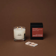 Load image into Gallery viewer, Ranger Station Cowboy Christmas candle with lit double wicks in a clear glass jar featuring a black cowboy hat illustration, displayed beside its black box with a red label reading &quot;amber, Montana juniper, frankincense, whiskey,&quot; and a small matchbox on a warm brown surface.
