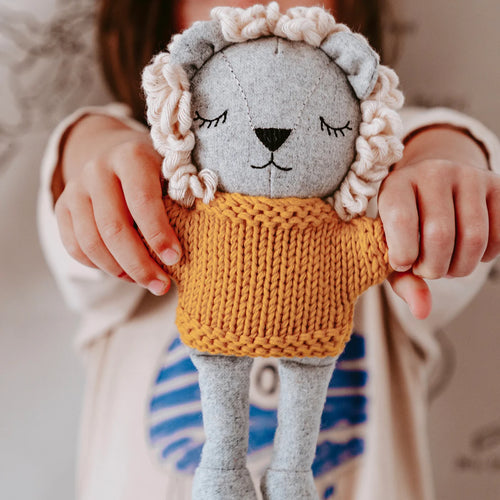 Plush merino-wool lion with a gray body, cream yarn mane, black embroidered facial features, and a mustard yellow knitted sweater, held upright by a child displayed against a blurred background. 