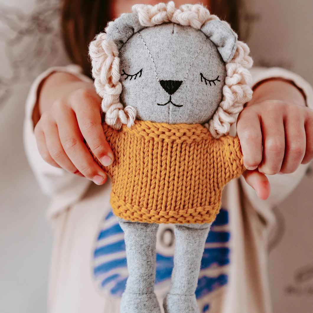 Plush merino-wool lion with a gray body, cream yarn mane, black embroidered facial features, and a mustard yellow knitted sweater, held upright by a child displayed against a blurred background. 