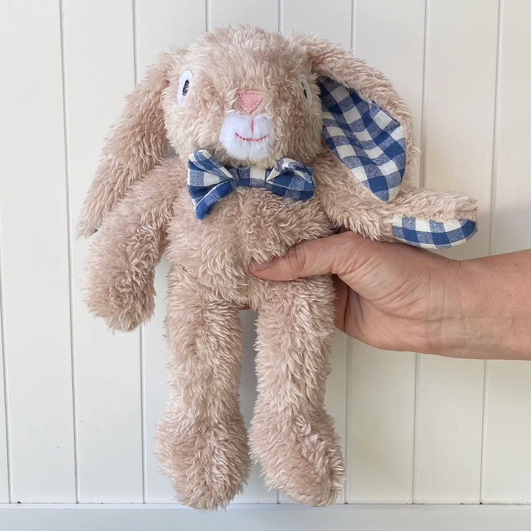 Vanilla-colored plush bunny with long floppy ears lined in blue and white gingham and a matching gingham bow tie, shown being held upright in front of a white paneled background. 