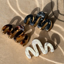Load image into Gallery viewer, Three wavy metal claw clips in tortoiseshell, black, and pearl displayed on a beige textured surface with soft curved light reflections. 