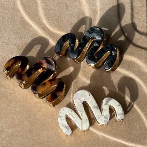 Three wavy metal claw clips in tortoiseshell, black, and pearl displayed on a beige textured surface with soft curved light reflections. 