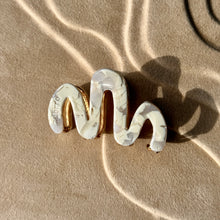 Load image into Gallery viewer, White wavy claw clip with gold hardware displayed on a beige textured surface with soft curved light reflections. 