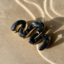Load image into Gallery viewer, Black wavy metal claw clip with gold hardware displayed on a beige textured surface with soft curved light reflections.