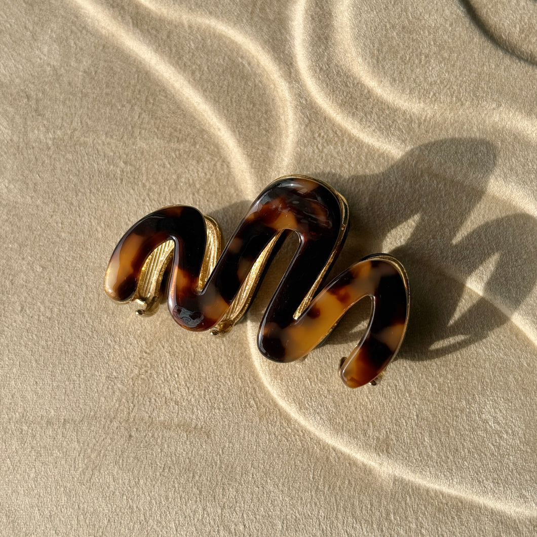 Tortoiseshell wavy metal claw clip with gold hardware displayed on a beige textured surface with soft curved light reflections. 