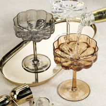 Load image into Gallery viewer, Amber, smokey, and clear handblown glass coupes with scalloped bowls styled on a gold tray, with champagne being poured into the amber glass, displayed on a soft white textured background. 